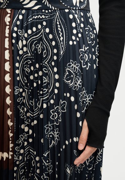 Πλισέ φούστα με σχέδια σε ναυτικό μπλε, καφέ και εκρού με λουλούδια και πουά, συνδυασμένη με μαύρο μακρυμάνικο τοπ με τρύπα για τον αντίχειρα.