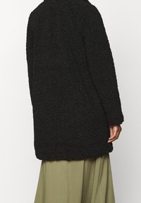 Cappotto lungo nero, textured e morbido, con vestibilità rilassata e maniche lunghe, abbinato a una gonna verde fluida, visto da dietro.