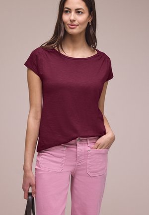 Vrouw met een bordeauxrode top met korte mouwen en lichtroze broek, met een hand in de zak en de andere hand die een tasgreep vasthoudt.