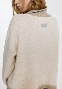 Cremefarbener, gestrickter Pullover mit einem gemütlichen Rollkragen, der einen dezenten Farbverlauf und ein Logoetikett auf der Rückseite aufweist. Weiche Textur und lockere Passform.