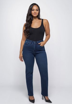 Vrouw met lang donker haar, gekleed in een zwart mouwloos topje, blauwe hoogwaterjeans en zwarte hakken, staand tegen een effen achtergrond.