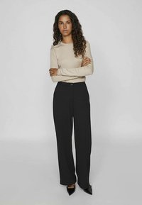 Bēša garām piedurknēm topi ar piegulošām piedurknēm, kombinēts ar melnām, platu jostasvietas biksēm un melnām augstpapēžu kurpēm ar smailiem purngaliem.