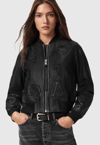 Chaqueta bomber de cuero negro con parches en relieve, cierre de cremallera, puños acanalados y bolsillos. Estilizada con un cinturón negro y jeans grises.