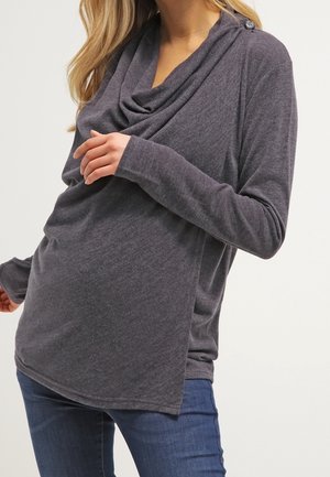 Vrouw die een losse donkergrijze top met lange mouwen en een gedrapeerde halslijn draagt, gecombineerd met een blauwe spijkerbroek, poserend met de ene hand bij de borst en de andere ontspannen langs het lichaam.