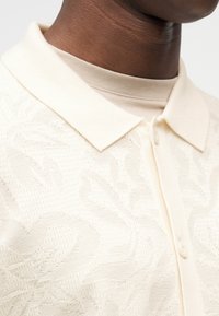 Μπεζ πόλο πουκάμισο με υφασμάτινο λουλουδάτο μοτίβο, που διαθέτει δομημένο γιακά, κλείσιμο με κουμπιά και στρώσεις υφάσματος από κάτω.