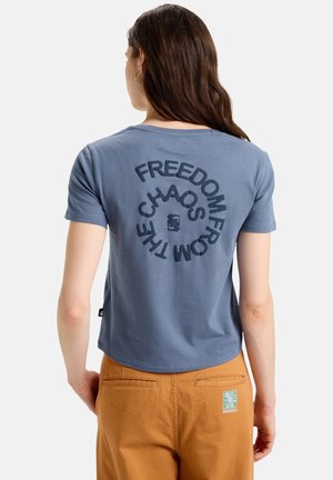 Blue cotton T-shirt with short sleeves and a rounded hem. Text on the back reads "FREEDOM FROM CHAOS." Worn with brown pants featuring a logo.