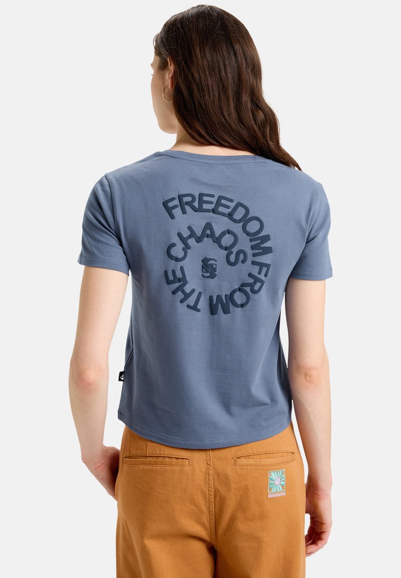 Blå bomullströja med korta ärmar och rundad nederkant. Texten på baksidan säger "FRIHET FRÅN KAOS." Buren med bruna byxor som har en logotyp.