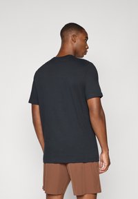 Black cotton t-shirt with short sleeves and crew neck, displayed from the back, paired with brown shorts. Simple design, no visible patterns.