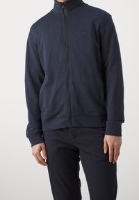 Marinblå zip-up sweatshirt med hög krage, ribbade muddar och nederkant. Har en liten logotyp på bröstet. Tillverkad av slät, texturerad tyg.