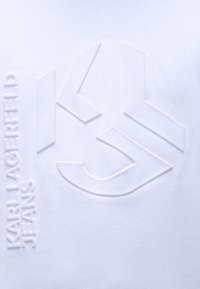 Balts kokvilnas T-krekls ar izcilu logotipu un tekstu "KARL LAGERFELD JEANS" kreisajā pusē. Gluda tekstūra, mūsdienīgs dizains.