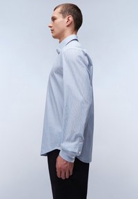 Lichtblauwe gestreepte shirt met witte en donkerblauwe verticale lijnen, knoopmanchetten, kraag en een relaxte pasvorm. Donkere broek.