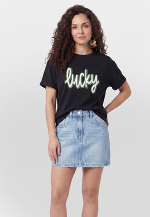 Donna con capelli ricci che indossa orecchini colorati con nappine, una t-shirt nera con il testo "lucky" e una gonna di denim azzurra, in piedi con le mani rilassate.