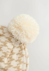 Stickad beige mössa med ett texturerat houndstooth-mönster, prydd med en fluffig vit pom-pom på toppen. Mjukt garnmaterial.