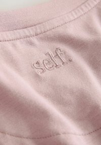 Svijetlopinkasta tkanina s glatkom teksturom. Ima izvezeni logo "self." blizu dekoltea. Detalji šavova vidljivi su na tkanini.