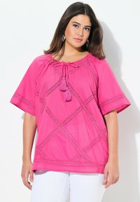 Top di cotone rosa con maniche corte, dettagli in pizzo con motivo a diamante e scollatura con fiocco e pon pon. Abbinato a pantaloni bianchi.
