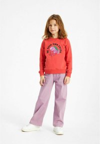 Röd sweatshirt med en paljett jordgubbe grafik och svart text, kombinerad med ljuslila vida byxor och vita sneakers.