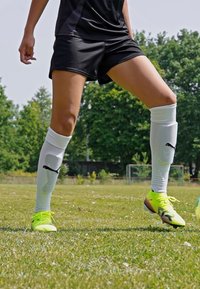 Shorts de sport noirs, chaussettes blanches à hauteur de genoux avec logo, et chaussures de football jaunes vives. Fond de terrain en herbe, journée ensoleillée, sans détails supplémentaires.