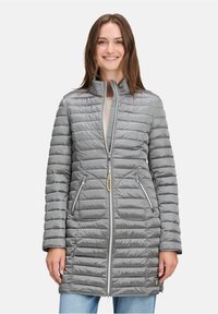 Smilende kvinde iført en lang grå quiltet jakke med to lynlåslommer og lynlås fortil, parret med lyseblå jeans.