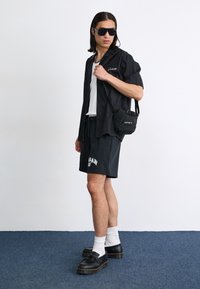 Black short-sleeve shirt over a white tank top, paired with black shorts featuring a logo, black bag, and chunky black shoes with thick soles.
