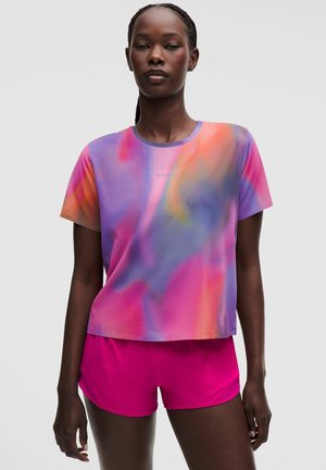 Mujer de pie con una camiseta multicolor pastel de efecto tie-dye y shorts deportivos rosa brillante sobre un fondo liso y claro.