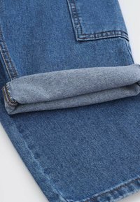 Jeans denimowe z podwiniętymi nogawkami, w średnim niebieskim kolorze, z widocznym szwem i teksturowaną powierzchnią materiału.