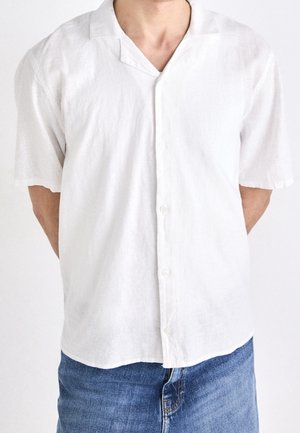 Mann trägt ein weißes Kurzarm-Button-up-Hemd mit lockerem Kragen und blaue Jeans, steht mit den Armen hinter dem Rücken.