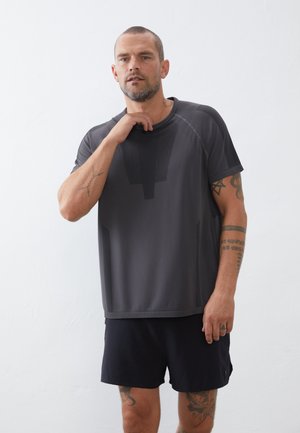 Dunkelgraues Sport-T-Shirt mit strukturierten Einsätzen und kurzen Ärmeln, kombiniert mit schwarzen Shorts. Zeigt Tattoos am Unterarm.