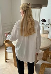 Túlméretezett fehér, hosszú ujjú ing, fekete nadrágra viselve. Simább textúra, laza szabás és hátul jéghát részlet.