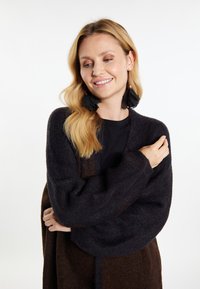 Tamna pletena cardigan s glatkom teksturom, s oversized rukavima i dizajnom u boji, uparena s velikim crnim pernatim naušnicama.