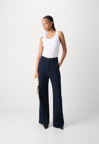 Navy high-waisted wide-leg trousers with textured fabric and front pockets, paired with a white sleeveless top and holding a small cream bag.