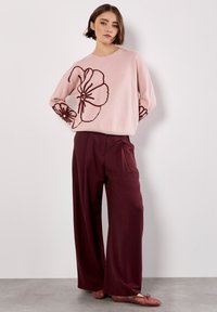 Lichtroze trui met donker bordeauxrode bloemenborduursels op de mouwen, gecombineerd met wijde bordeauxrode pantalon en roze ballerina's. Zachte textuur.