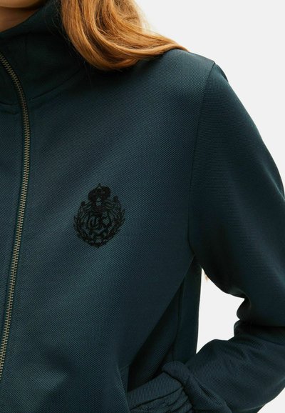 Donkergroen sportjacket met een hoge kraag, gestructureerde stof, gouden rits en een zwart geborduurd logo op de linkerborst.