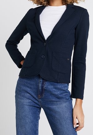 Person trägt einen engen schwarzen Blazer über einem weißen Hemd mit blauen, hoch taillierten Jeans und posiert mit einer Hand auf der Hüfte und der anderen entspannt.