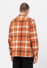 Camisa de franela a cuadros en naranja y blanco con acentos marrones, presenta mangas largas y un cuello abotonado, hecha de una tela texturada.