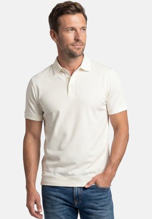 Mężczyzna o krótkich brązowych włosach, ubrany w biały polo z krótkim rękawem i niebieskie dżinsy, stoi z jedną ręką w kieszeni, patrząc w prawo.