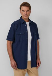 Korte mouwen marineblauwe overhemd met knopen, gemaakt van zachte stof, met twee voorkruiken en lichtblauwe knopaccenten. Draagt over een wit T-shirt.
