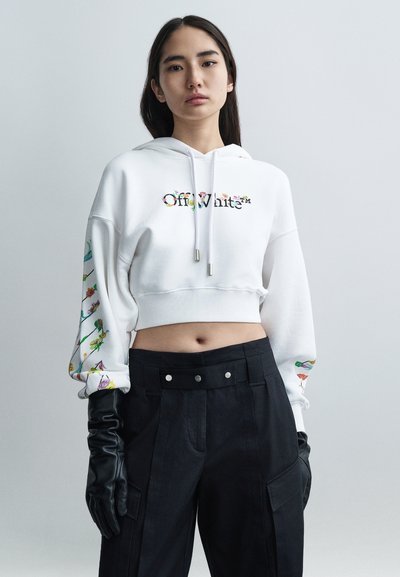 Hoodie blanc court avec broderie florale multicolore, doté d'un cordon de serrage et de détails en métal argenté. Porté avec un pantalon noir taille haute et des gants noirs.