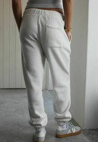 Isik, kes kannab valgeid spordipükse tagataskuga, halli särkiga, valgete sokkide ja heledate hallide tossudega, seisab siseruumides betoonpinnal.