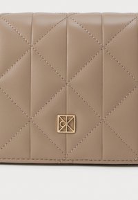 Bežs kviltots ādas maciņš ar diognālu metienu, priekšā ar nelielu zelta ģeometrisku logo akcentu.