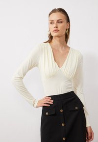Long-sleeve, ivory bodysuit with a deep V-neck and gathered detail at the bust, paired with a high-waisted black skirt featuring gold buttons.