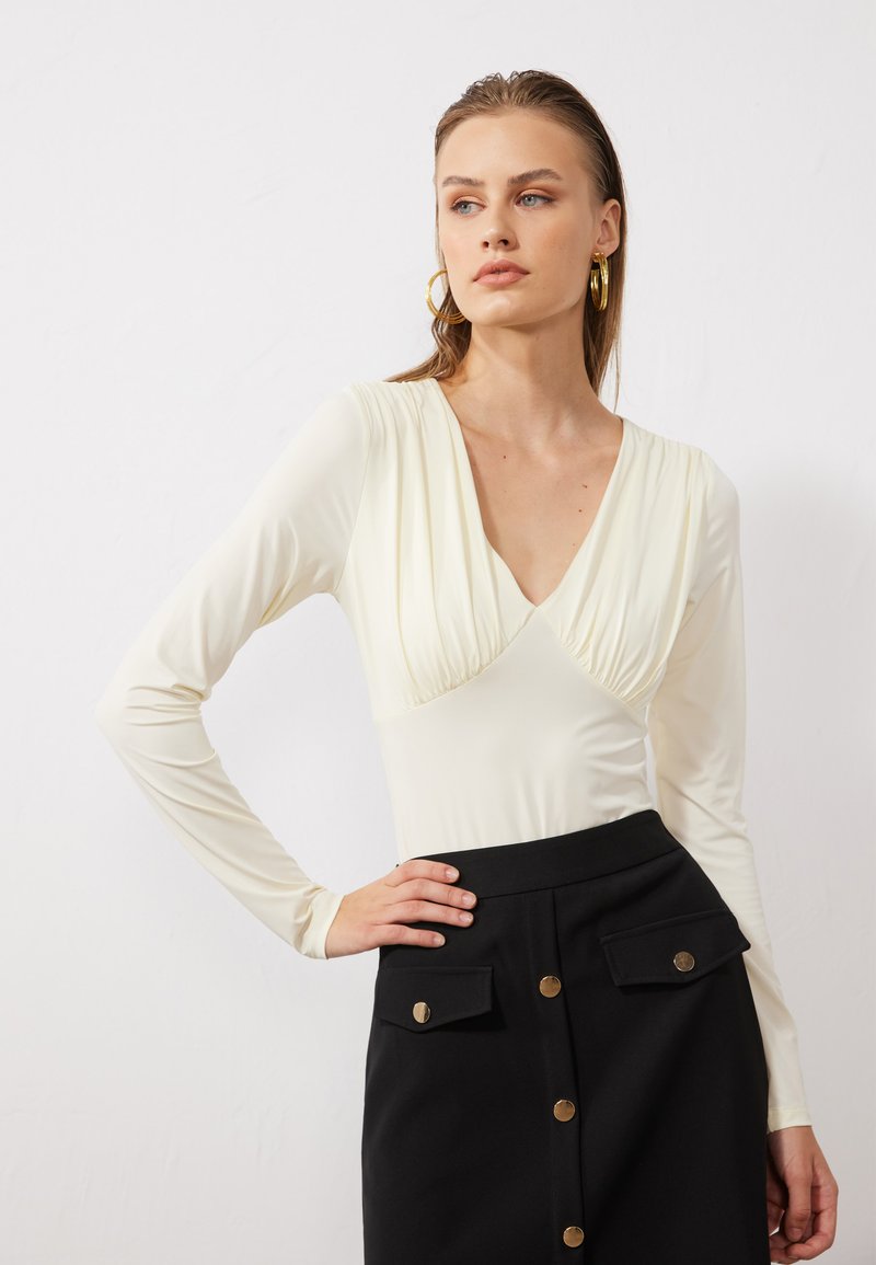 Long-sleeve, ivory bodysuit with a deep V-neck and gathered detail at the bust, paired with a high-waisted black skirt featuring gold buttons.