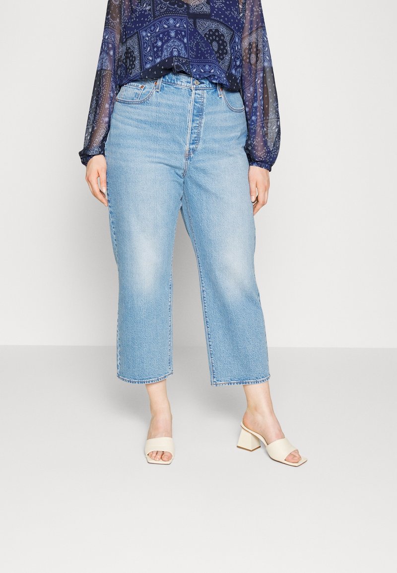 Femme portant un jean court bleu clair, un chemisier transparent bleu marine à motifs, et des sandales crème à talon bloc et bouts ouverts, debout sur un sol gris.
