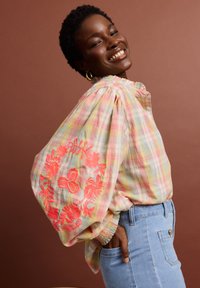 Personne souriante portant un chemisier à carreaux pastel avec des manches bouffantes et une broderie florale corail vif, associée à un jean bleu clair sur un fond marron.