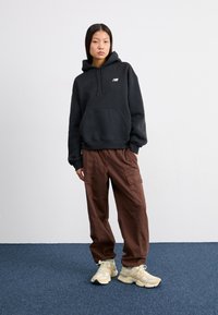 Sweatshirt noir à capuche avec poche avant, associé à un pantalon cargo marron ample et des baskets beiges avec des accents crème et des lacets.