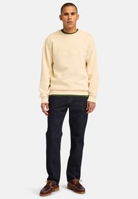 Sweatshirt crème avec logo "Timberland" en relief, col rond vert, jean sombre et chaussures bateau bordeaux avec semelles beiges.