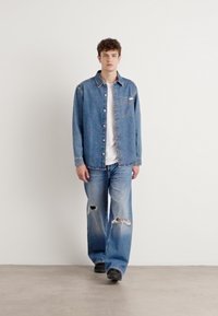 Camisa de mezclilla azul con bolsillo frontal, diseño abotonado, llevada sobre una camiseta blanca. Combinada con jeans de pierna ancha de lavado claro y desgastados, y zapatos negros.