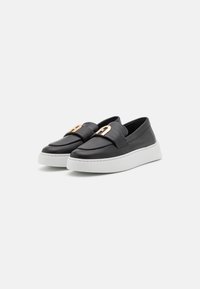 Mocasines de cuero negro sin cordones con suelas de goma blanca gruesa y hebillas decorativas de metal dorado en la parte superior, mostrados uno al lado del otro sobre un fondo blanco.