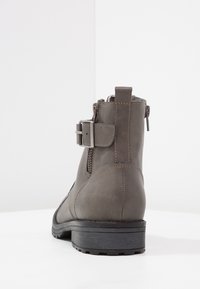 Šedá kožená kotníková bota s bočním zipem, nastavitelným popruhem a texturovaným povrchem. Pevná černá gumová podrážka s protiskluzovým vzorem.