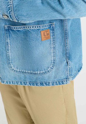 Lichtblauwe spijkerjassenzak met bruine leren logo patch op beige broek tegen een effen achtergrond.