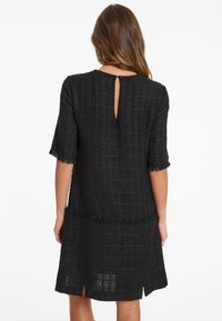 Schwarzes Kleid aus strukturiertem Stoff mit einem Gittermuster, kurzen Ärmeln und einem Schlüsselaugenöffnung auf der Rückseite mit einem Knopf. Fransenbesatz am Saum.